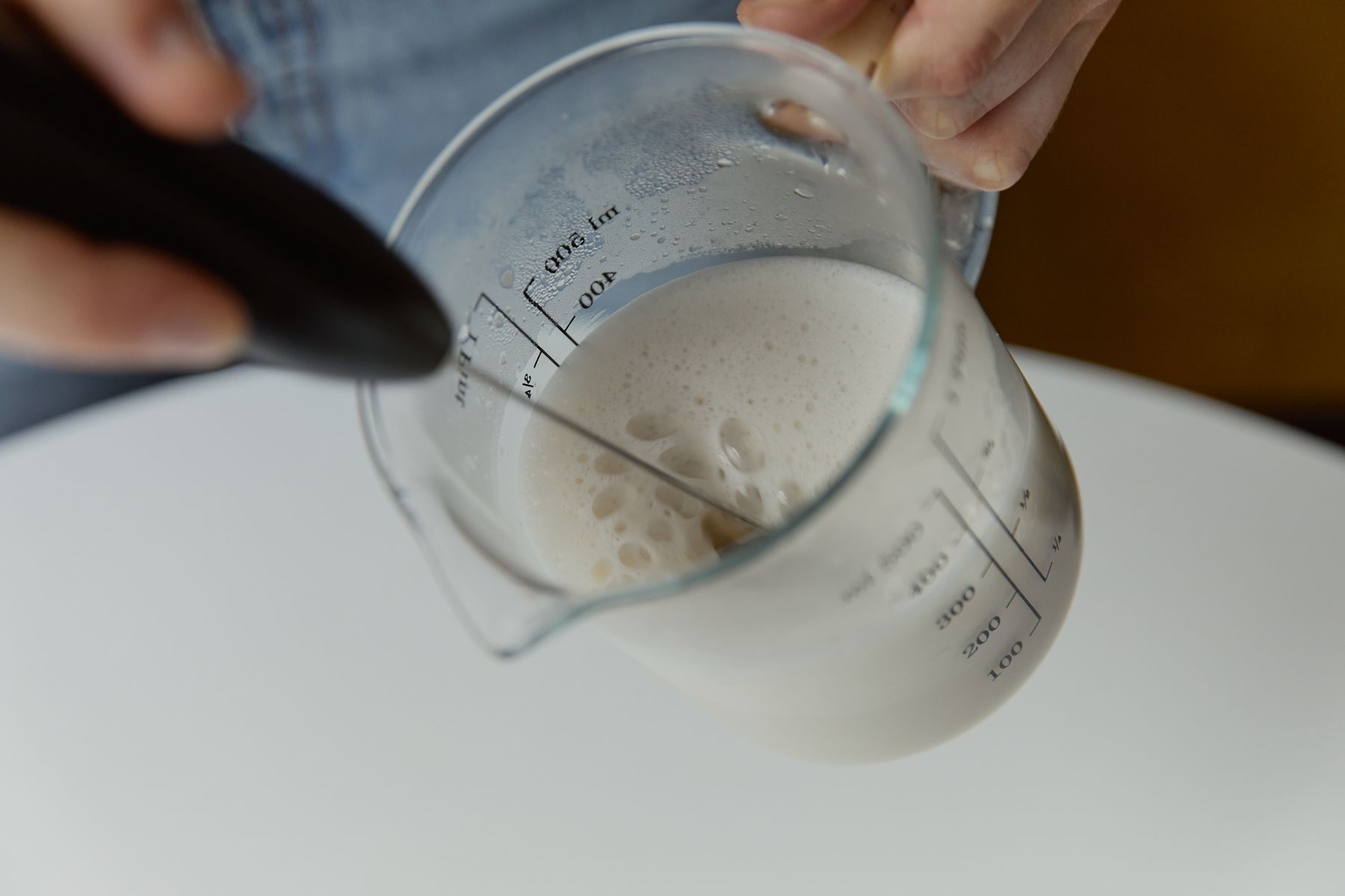 Hand hält Milchaufschäumer in Messbecher mit frisch aufgeschäumter Milch