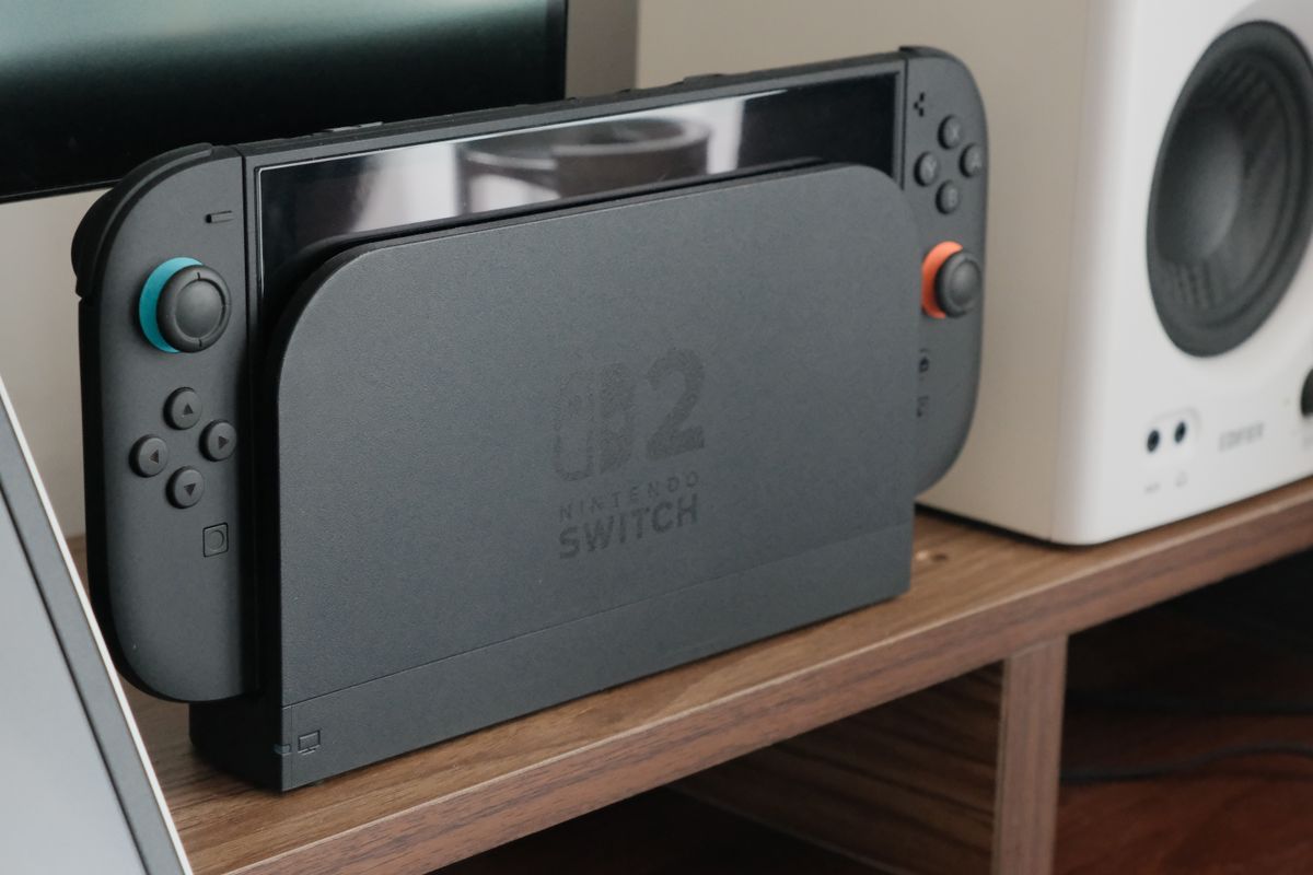 Nintendo Switch 2 steckt in der Dockingstation auf einem Holzregal neben einem Lautsprecher