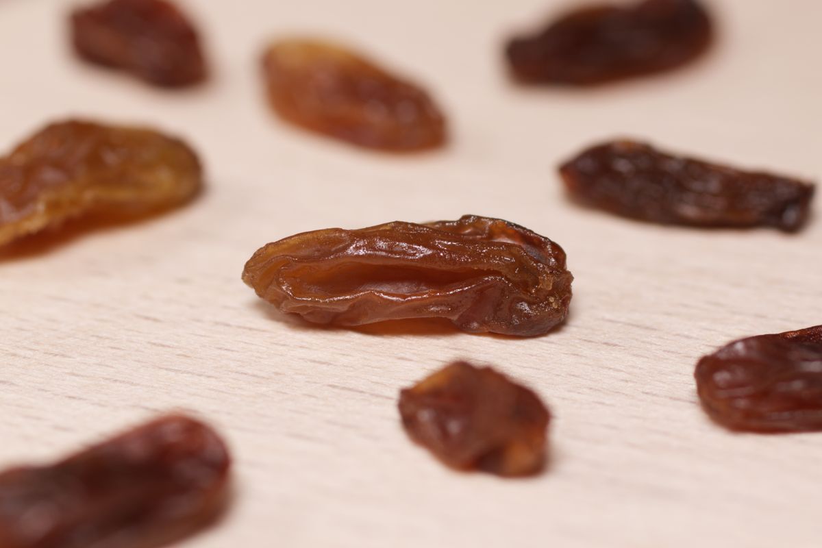 Einzelne Rosinen liegen verstreut auf einer hellen Holzfläche, eine davon groß im Vordergrund