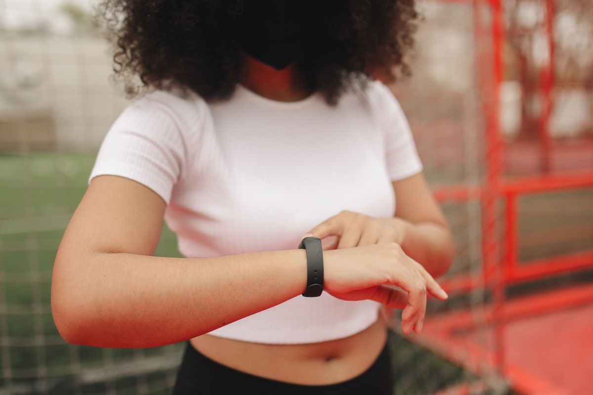 Person im weißen Shirt bedient auf einem Sportplatz mit dem Finger ein schmales schwarzes Fitnessarmband am linken Handgelenk