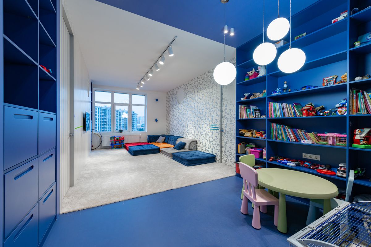 Spielzimmer mit blauen Wandregalen voller Bücher und Spielsachen, davor ein kleiner Tisch mit Kinderstühlen